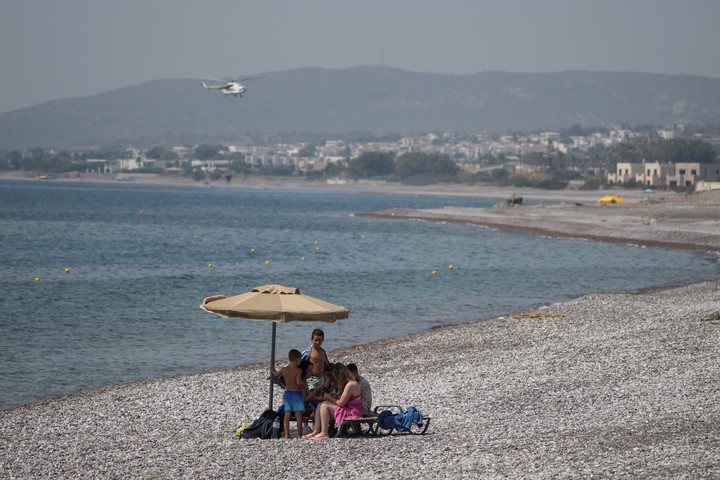 La gente se siente bajo la sombra mientras un helicóptero sobrevuela durante un incendio forestal en una playa cerca del pueblo de Gennadi, en la isla de Rodas, en el Mar Egeo, en la seguridad de Grecia.  (Foto AP/Petros Giannakouris)